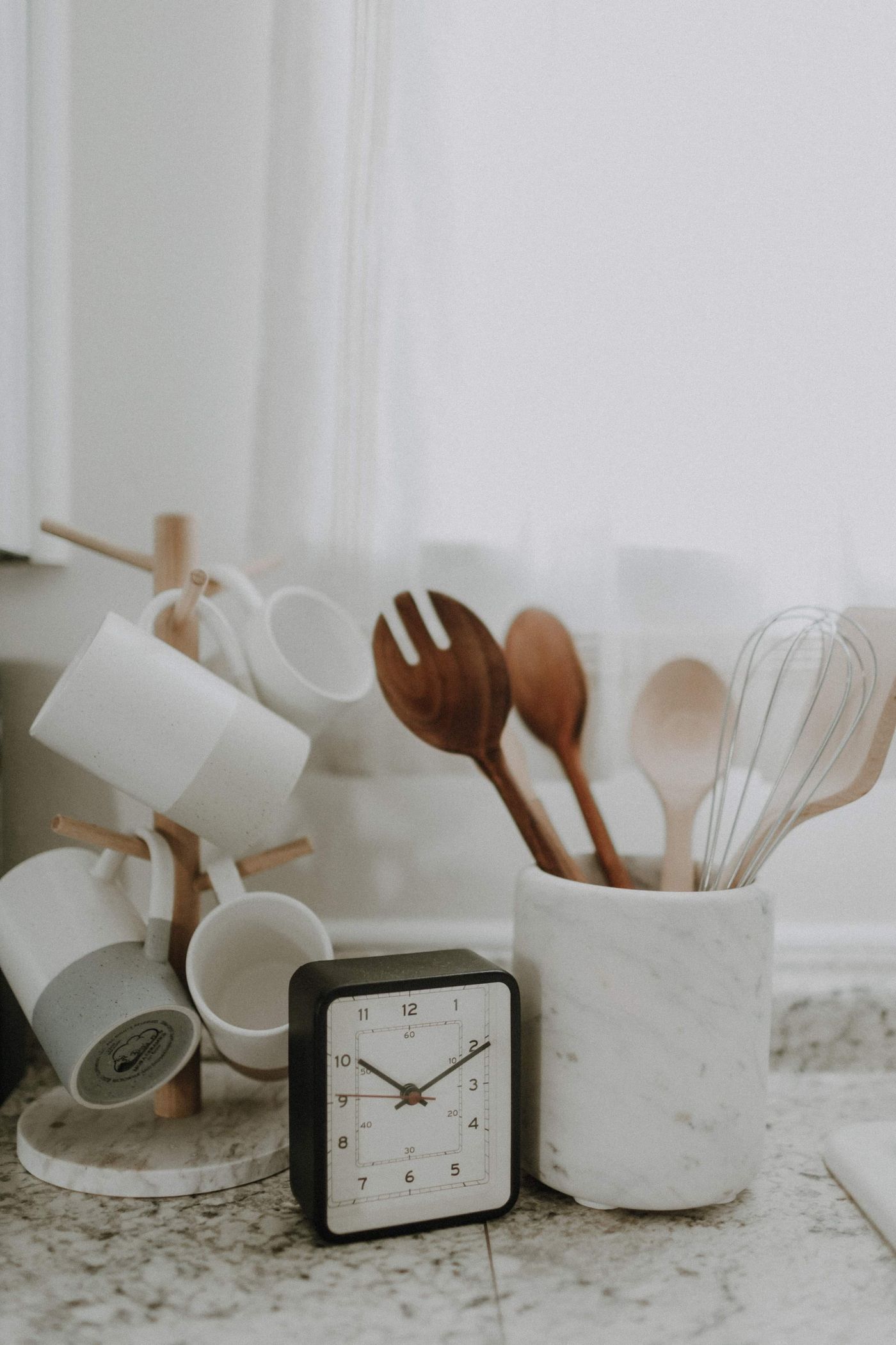 A small clock beside mugs and utensils on a kitchen counter