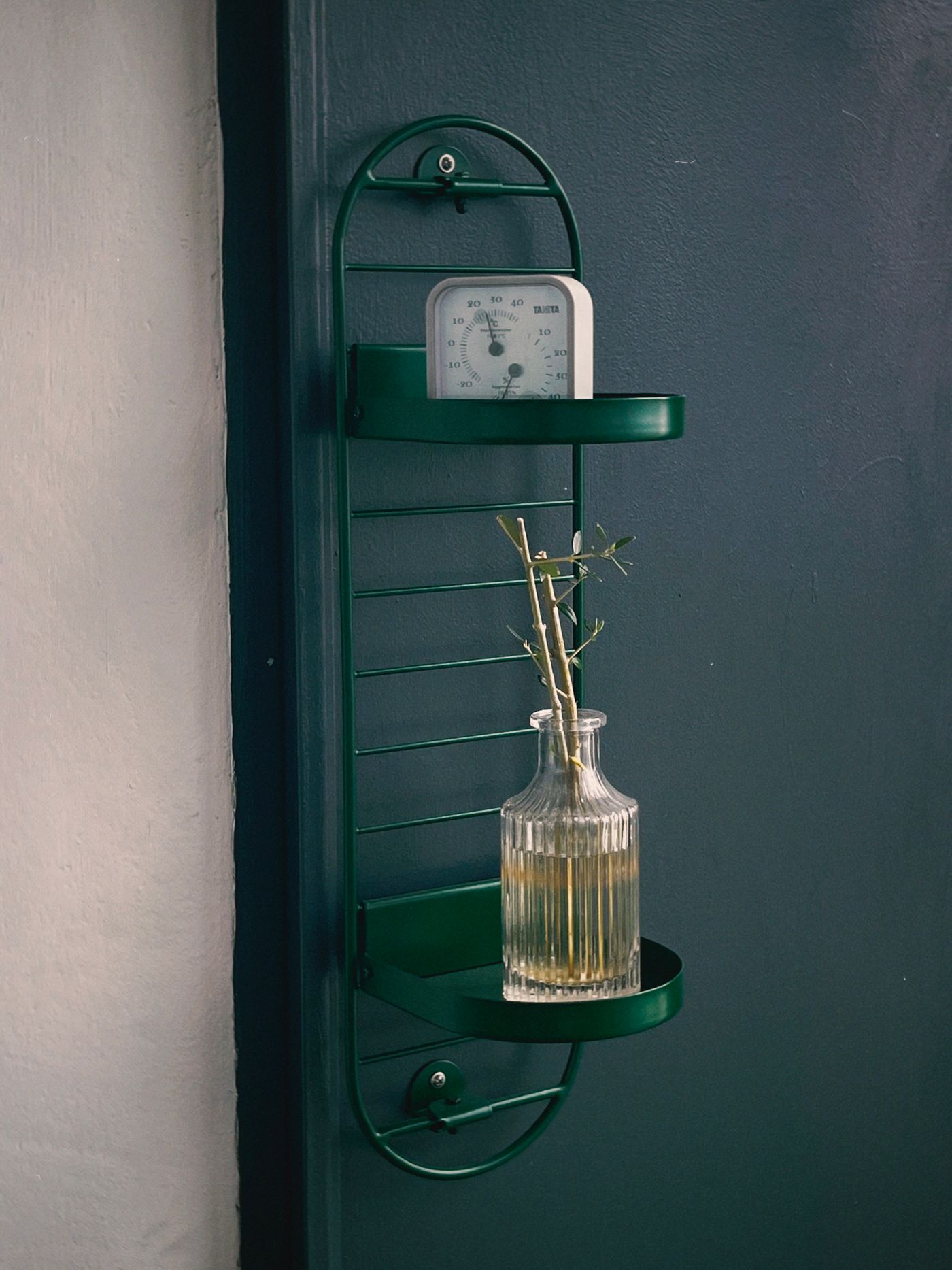 A small square clock on an open metal wall shelf above a glass vase